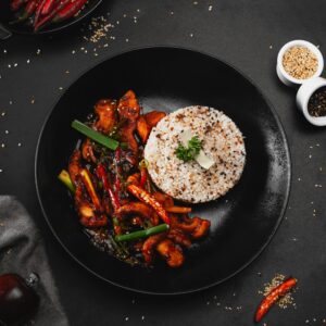 A tantalizing Asian stir-fry with chicken and vegetables beside seasoned rice on a black plate.
